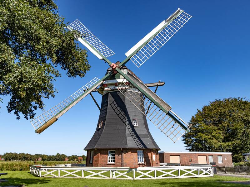 Man kann hier die Tjaden Mühle sehen. Sie ist grau und um ihr rumzu steht ein weißer, kleiner Zaun.